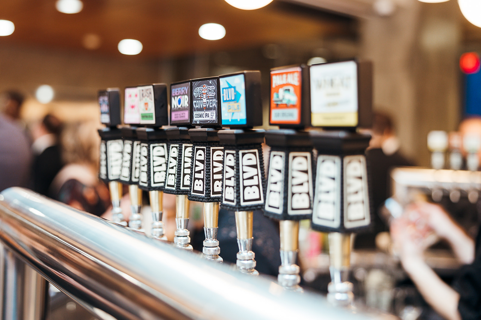 A row of beer taps with various labels in a bar setting. The handles display different brand names and logos, with a blurred background showing patrons and bar details, creating a lively atmosphere.