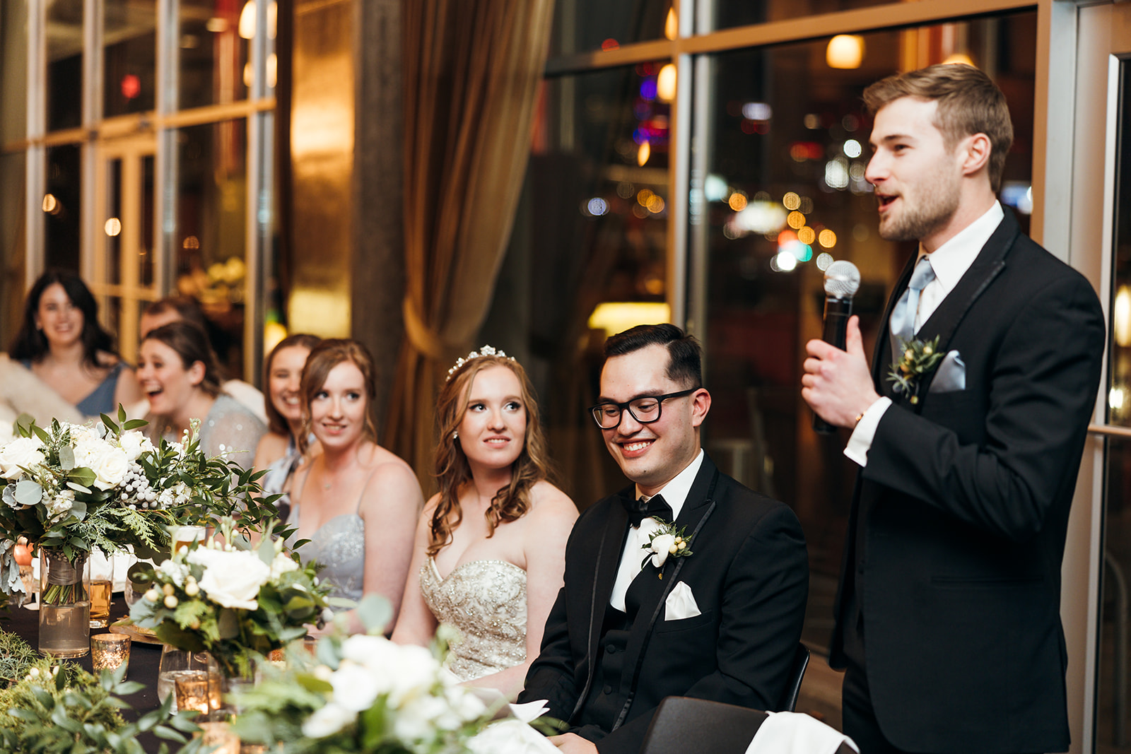 A man in a suit gives a speech with a microphone at a wedding reception. The bride and groom, smiling, sit at a table adorned with flowers and candles. Bridesmaids are seated nearby, listening attentively.