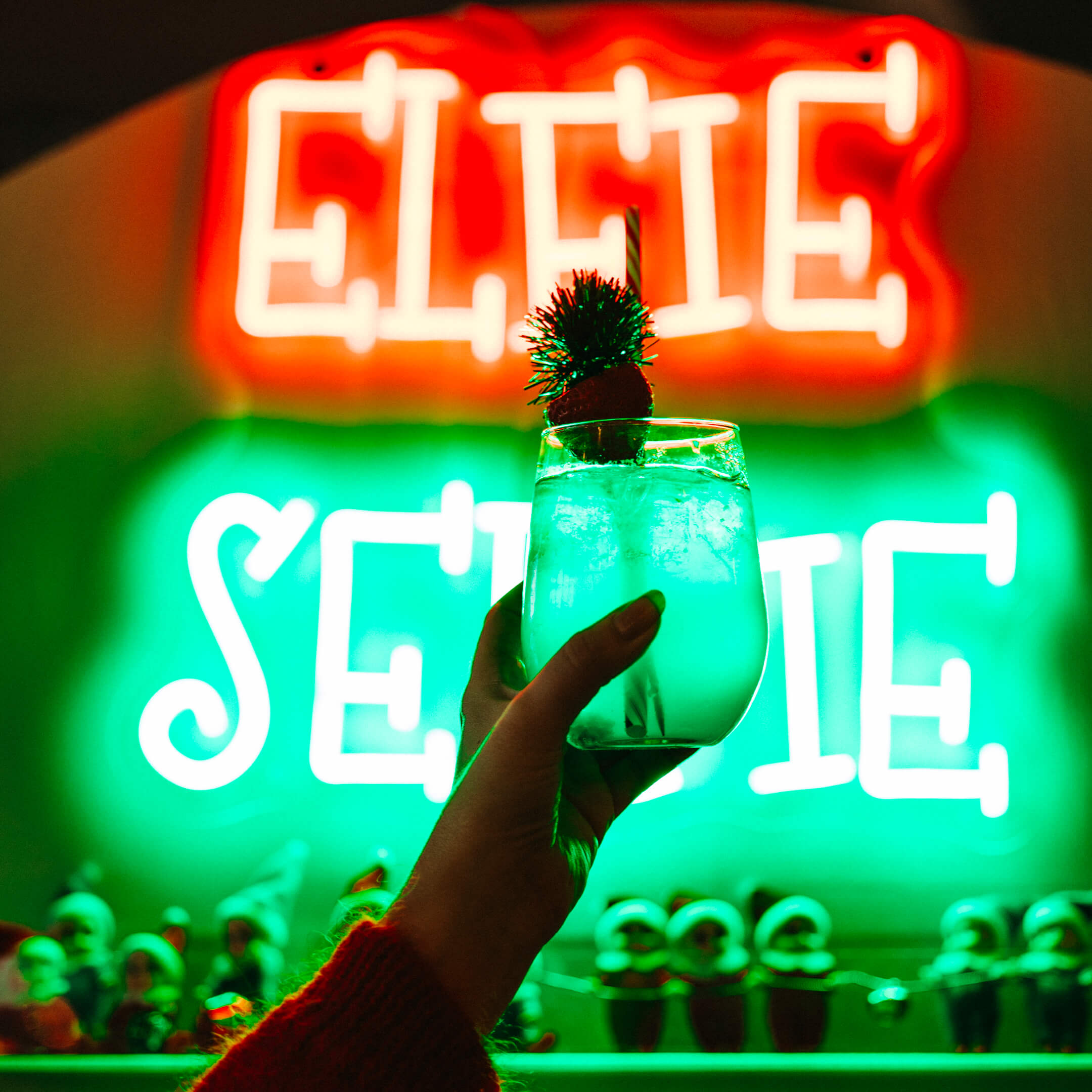 A hand holding a cocktail garnished with a small pom-pom. In the background, there are festive neon lights in red and green spelling "ELFIE SELFIE," with miniature elf figures lined up below.