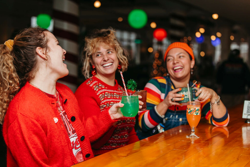 Three people sitting at a wooden bar, laughing and holding colorful drinks. They wear festive clothing in red, orange, and multicolored patterns. The setting features holiday decorations with green and red lights.