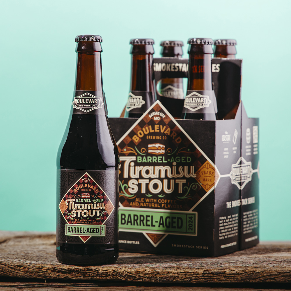 A bottle and a six-pack of Boulevard Brewing Co.'s Barrel-Aged Tiramisu Stout are placed on a wooden surface with a turquoise background. The label highlights coffee and natural flavors.