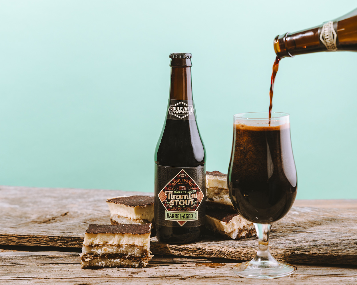 A bottle of Boulevard Brewing Co. Barrel-Aged Imperial Stout is next to a glass being filled with dark beer. Three cream-topped chocolate brownies are placed nearby on a wooden surface, set against a light turquoise background.