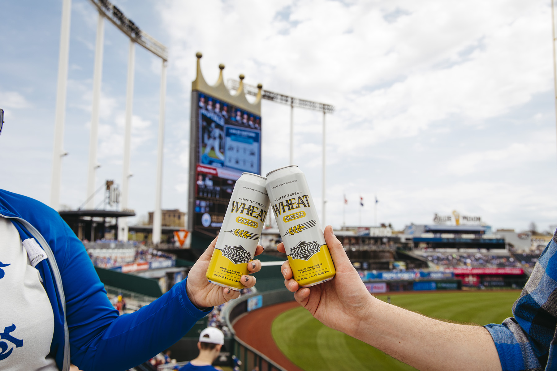 Two friends excitedly hold up cans of wheat beer at a bustling baseball stadium, the vibrant field and towering scoreboard visible in the background. The sky is partly cloudy, adding to the lively atmosphere buzzing with anticipation about the game.