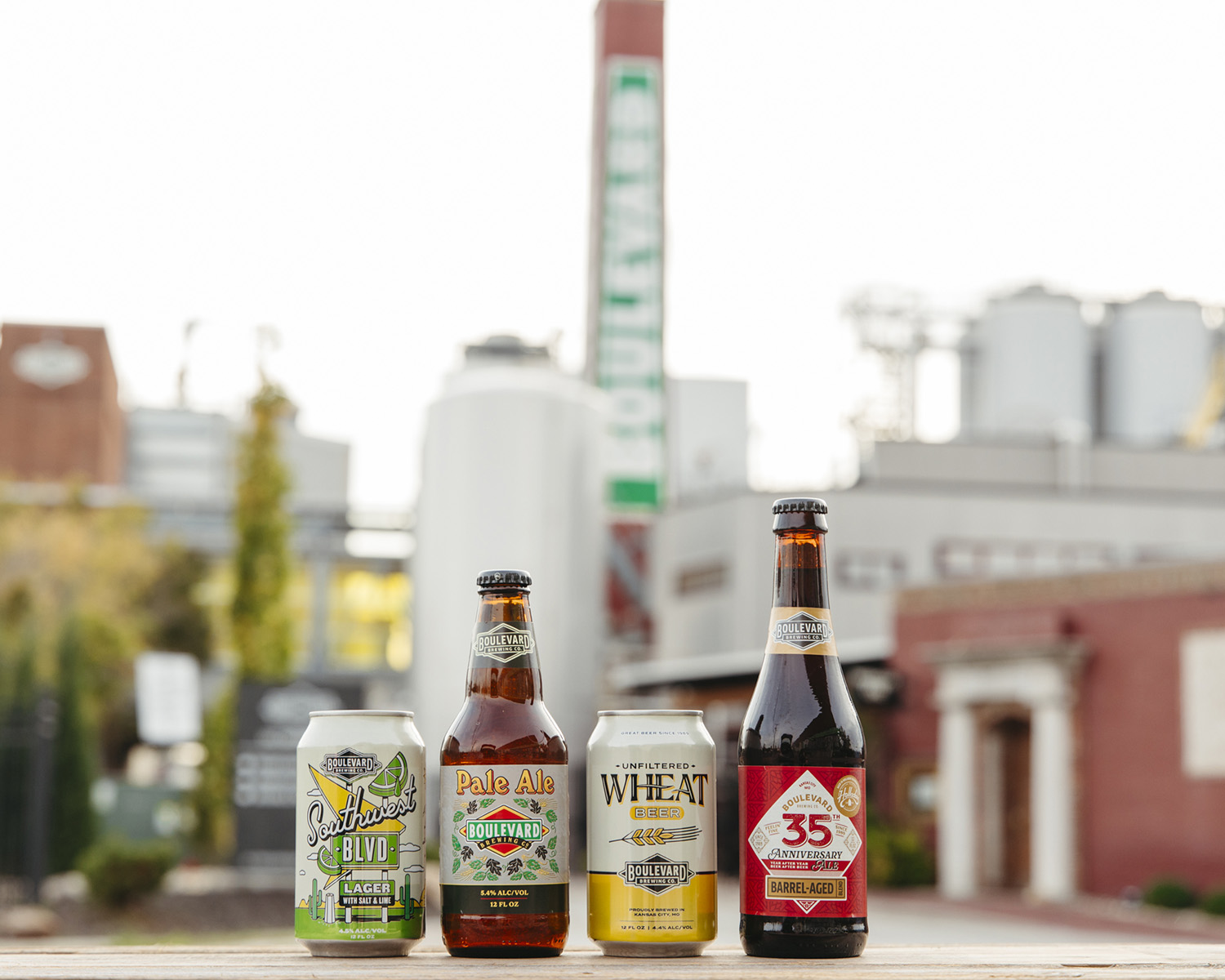 Four Boulevard Brewing Company beer bottles and cans are displayed on a wooden surface. The backdrop features the brewery's industrial buildings, including a tall chimney with the Boulevard logo. The setting conveys a craft beer atmosphere.