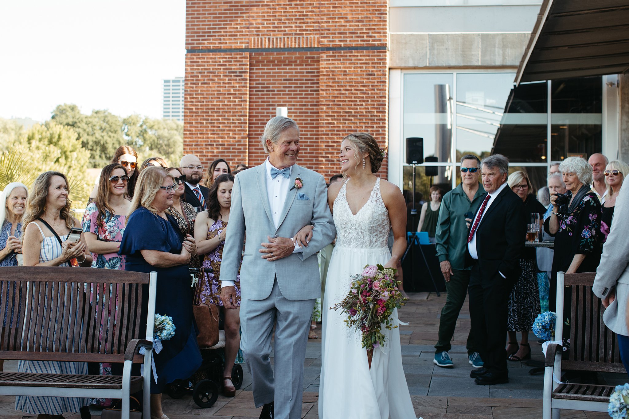 A bride and a man, both smiling, walk down an outdoor aisle lined with seated and standing guests. The bride holds a bouquet and wears a white gown, while the man is in a light gray suit. The background features a brick building and greenery.