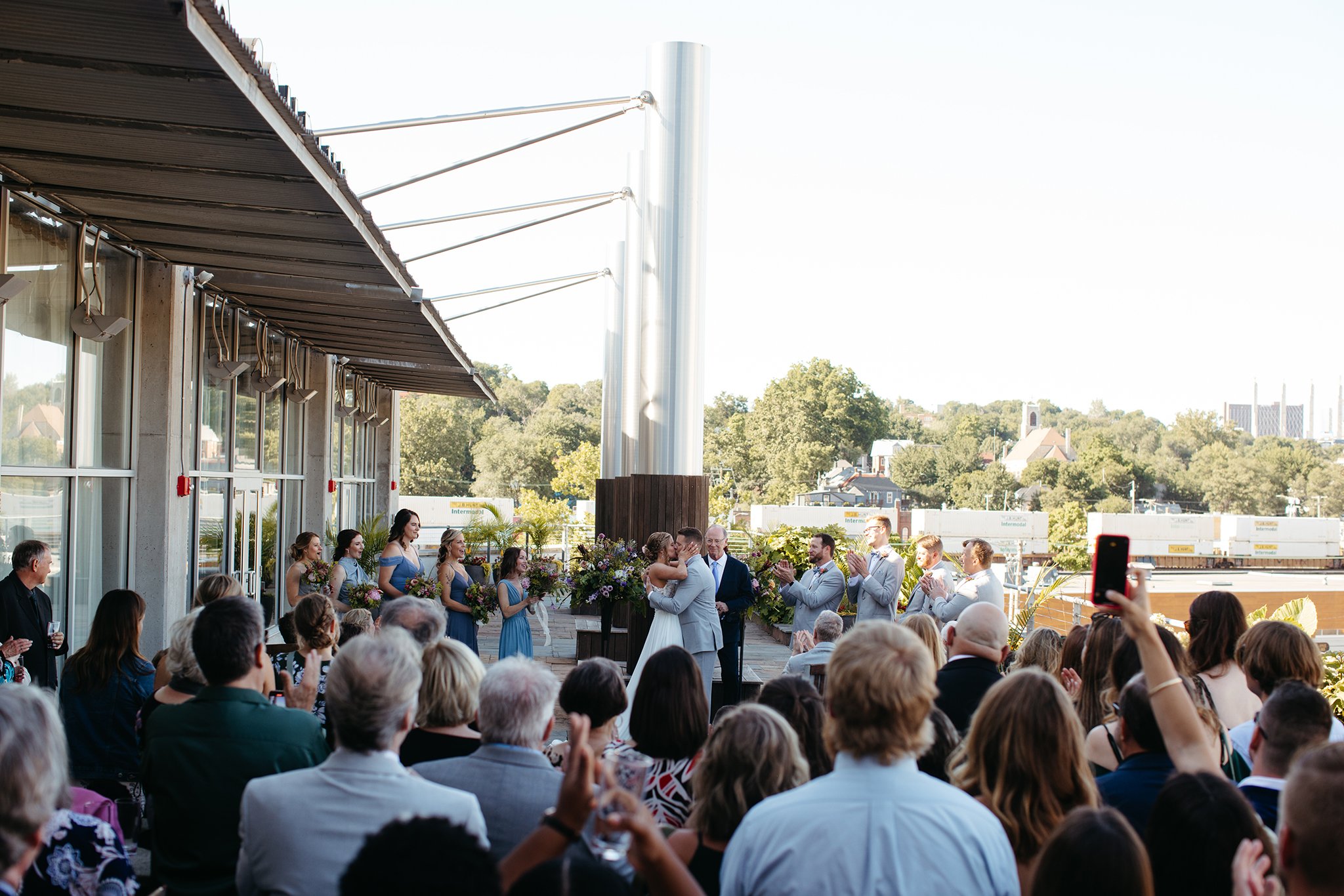 A wedding ceremony on an outdoor terrace with a large audience. The couple is kissing at the altar, surrounded by their wedding party holding flowers. Guests are seated and some are standing, capturing the moment with phones. Trees are visible in the background.