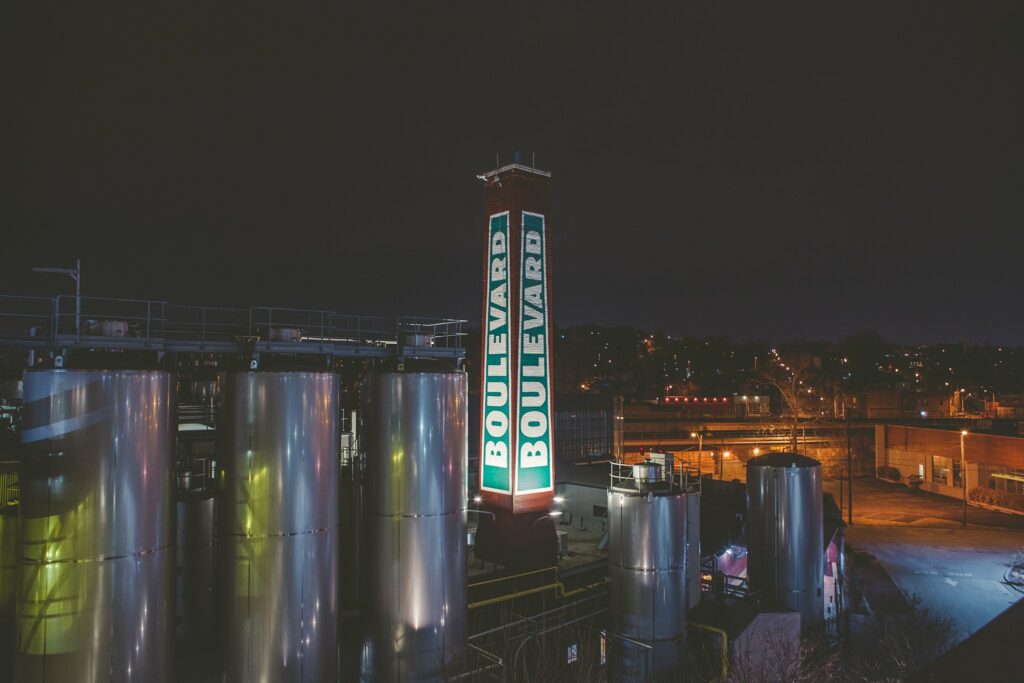 Night view of an industrial facility with large metal tanks and a tall illuminated chimney displaying the word "Boulevard" repeatedly. City lights are visible in the background.