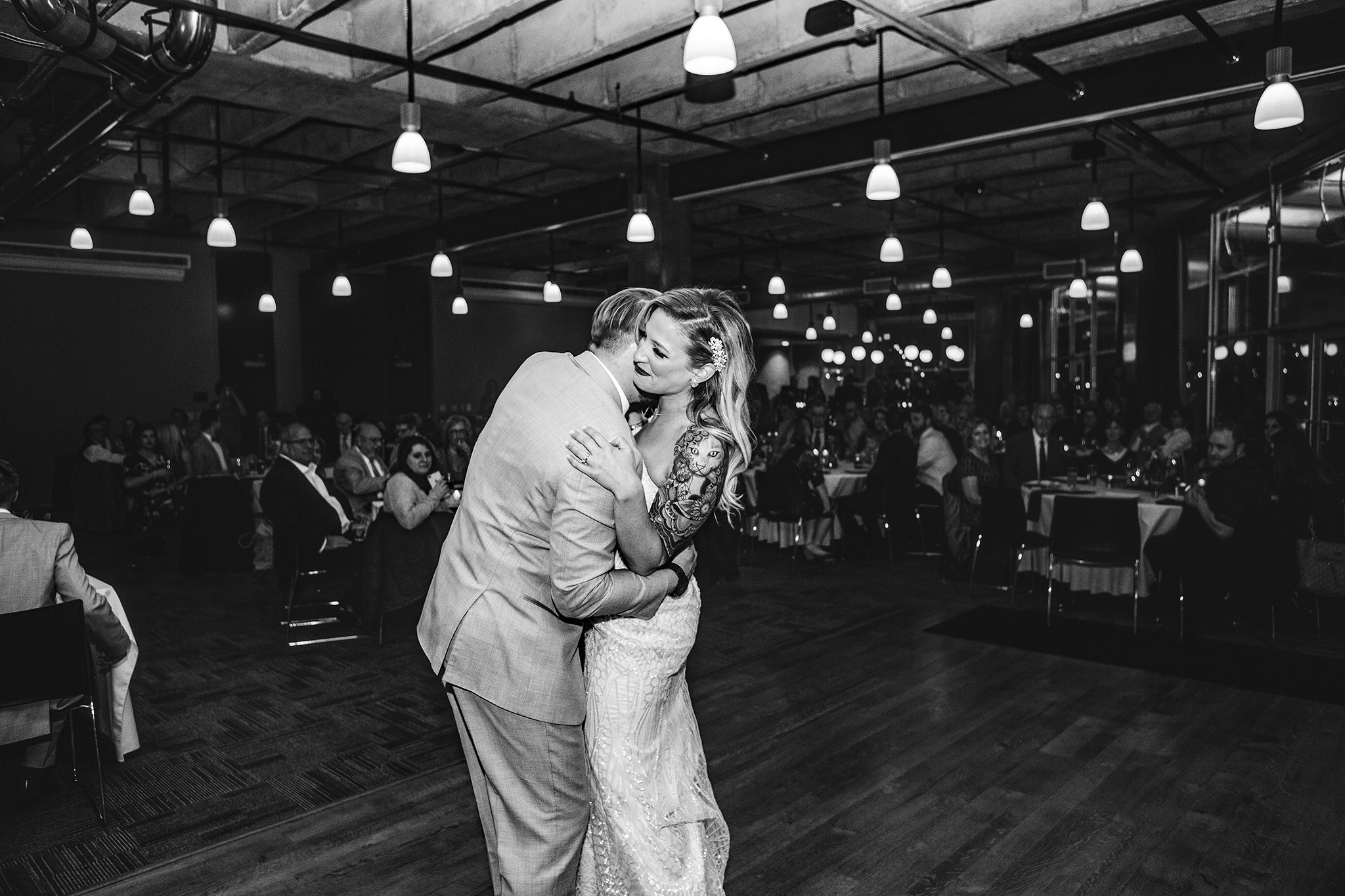 A black-and-white photo of a bride and groom sharing a joyful dance in a warmly lit reception hall, surrounded by seated guests. The bride wears a lace gown and the groom is in a suit. Hanging lights create a festive atmosphere.