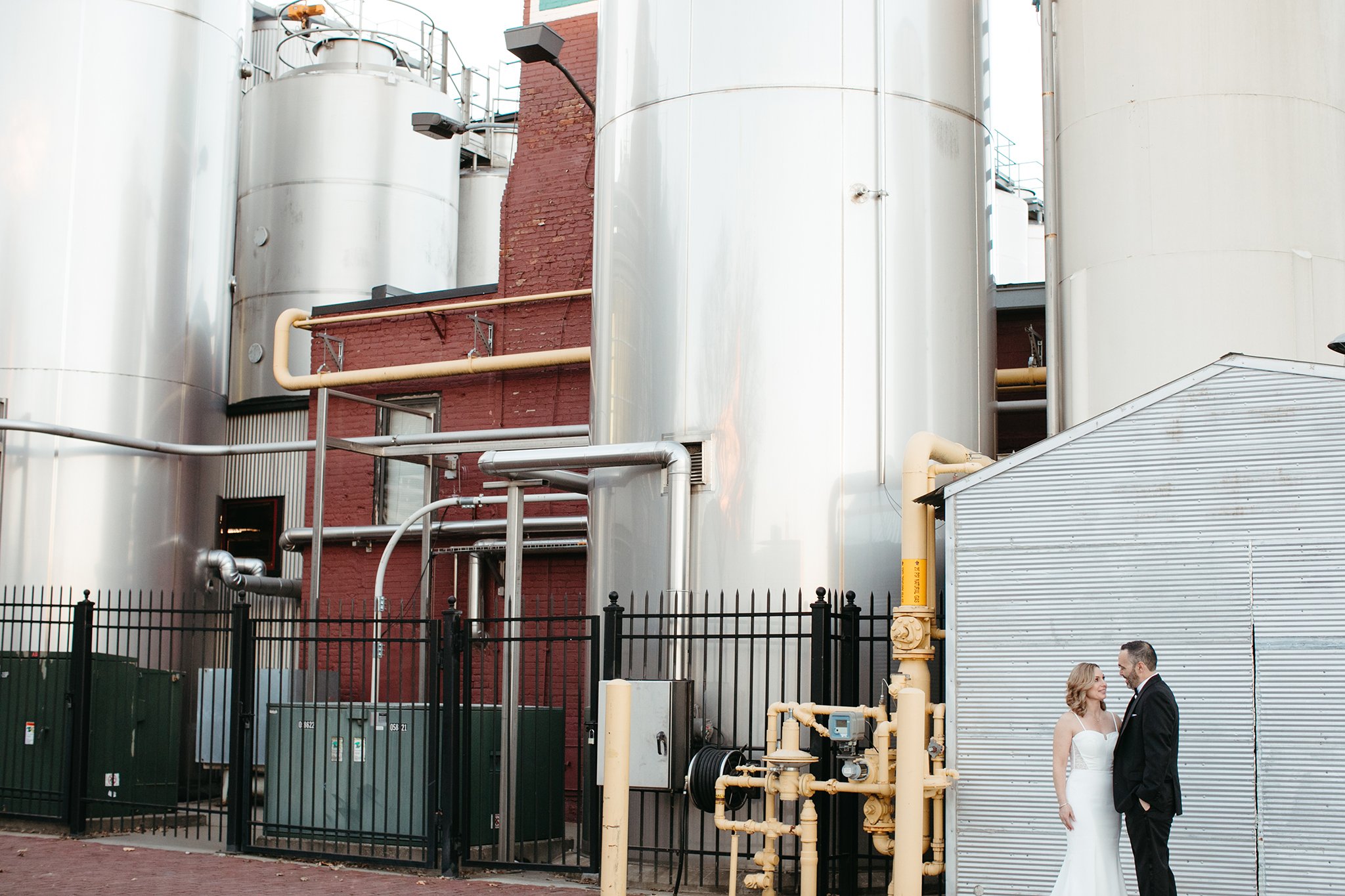 A couple in wedding attire stands in an industrial setting with large metal tanks and a brick wall in the background. They gaze at each other, surrounded by industrial pipes and fenced equipment.