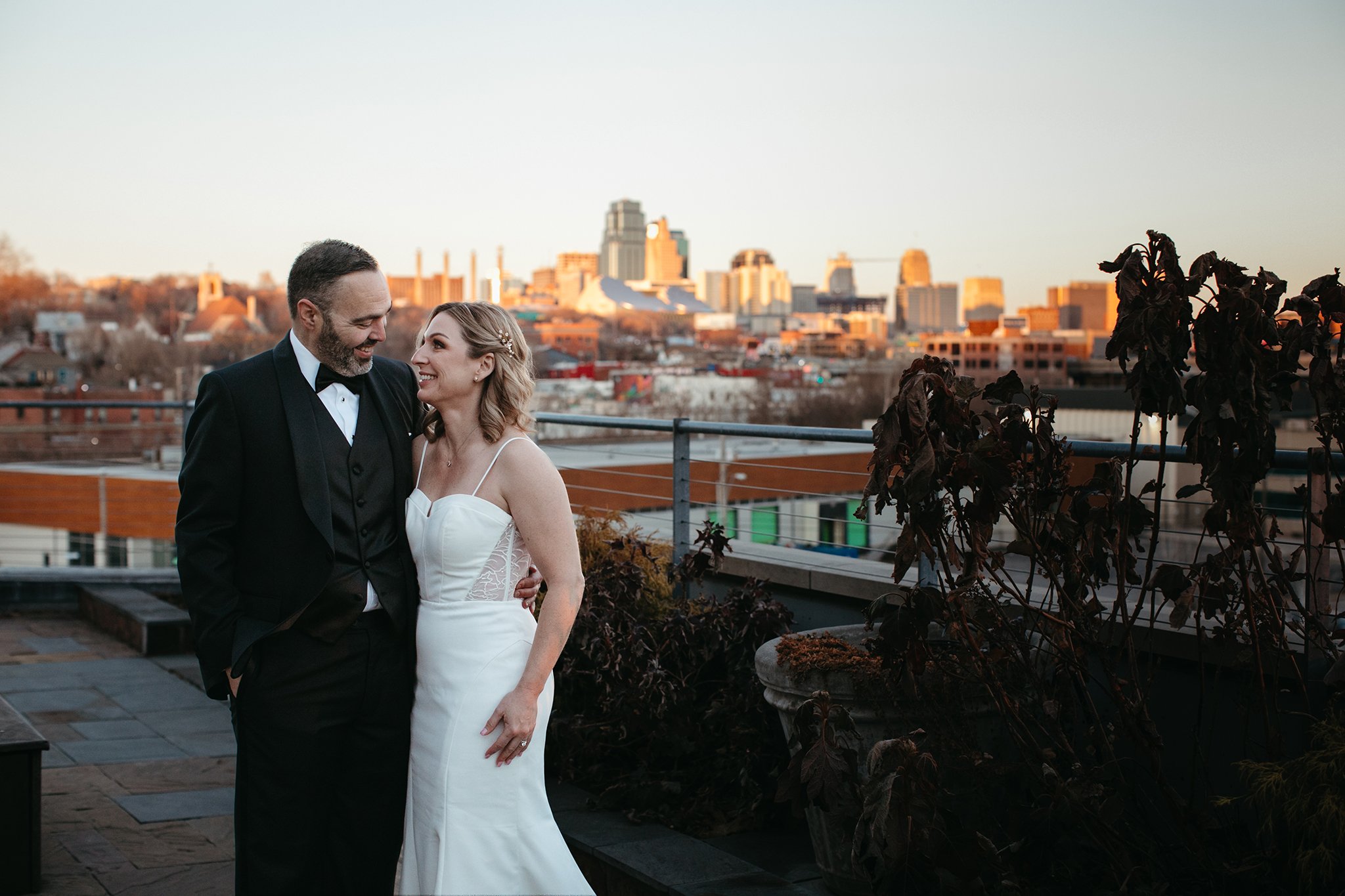 A bride and groom share a joyful moment on a rooftop, with a city skyline in the background. The bride wears a white gown, and the groom a dark suit. The sun sets, casting a warm glow over the scene.