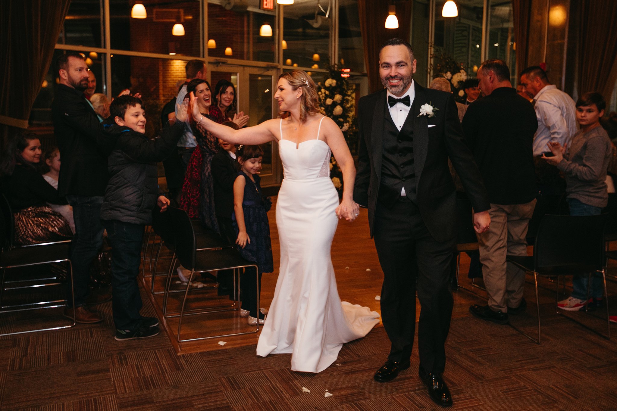 A bride and groom walk down the aisle smiling and holding hands. The bride is high-fiving a young boy in the crowd, while guests applaud and celebrate. The setting is warmly lit and festive.