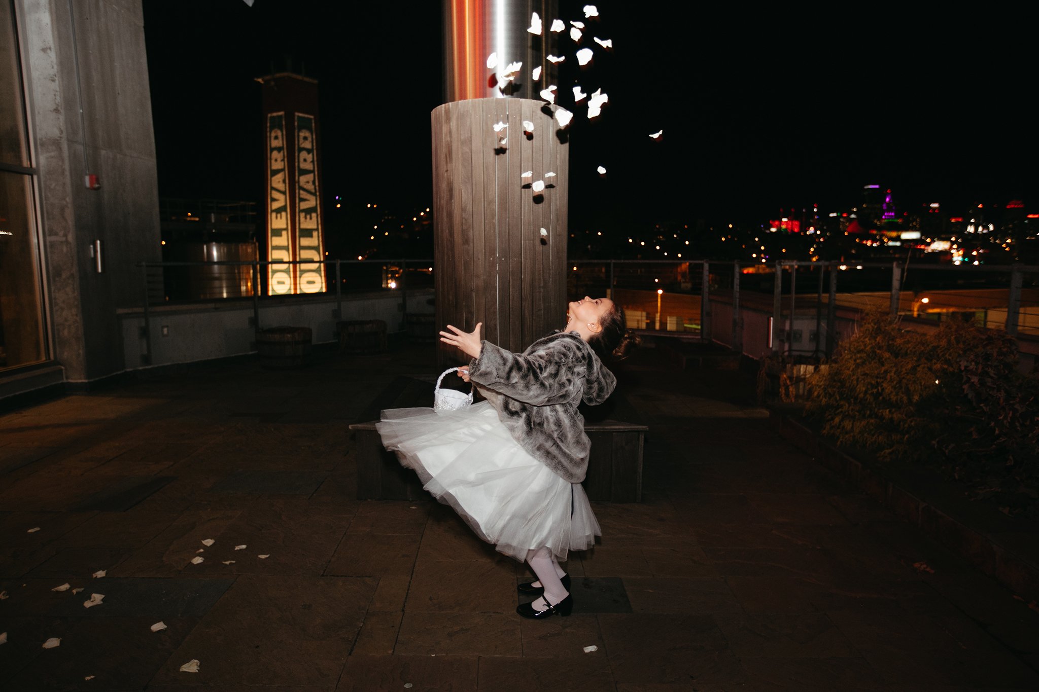 A young child in a white dress and a fur coat joyfully tosses rose petals into the air at night. City lights and a sign reading "B O U L E V A R D" illuminate the background.