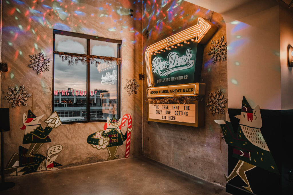 A festive indoor scene at Rec Deck the Halls sparkles with colorful lights reflecting on the walls. Signs proclaim "Good Times, Great Beer," while holiday-themed cutouts of elves and candy canes add charm. A window reveals a cityscape backdrop, completing the cozy ambiance.