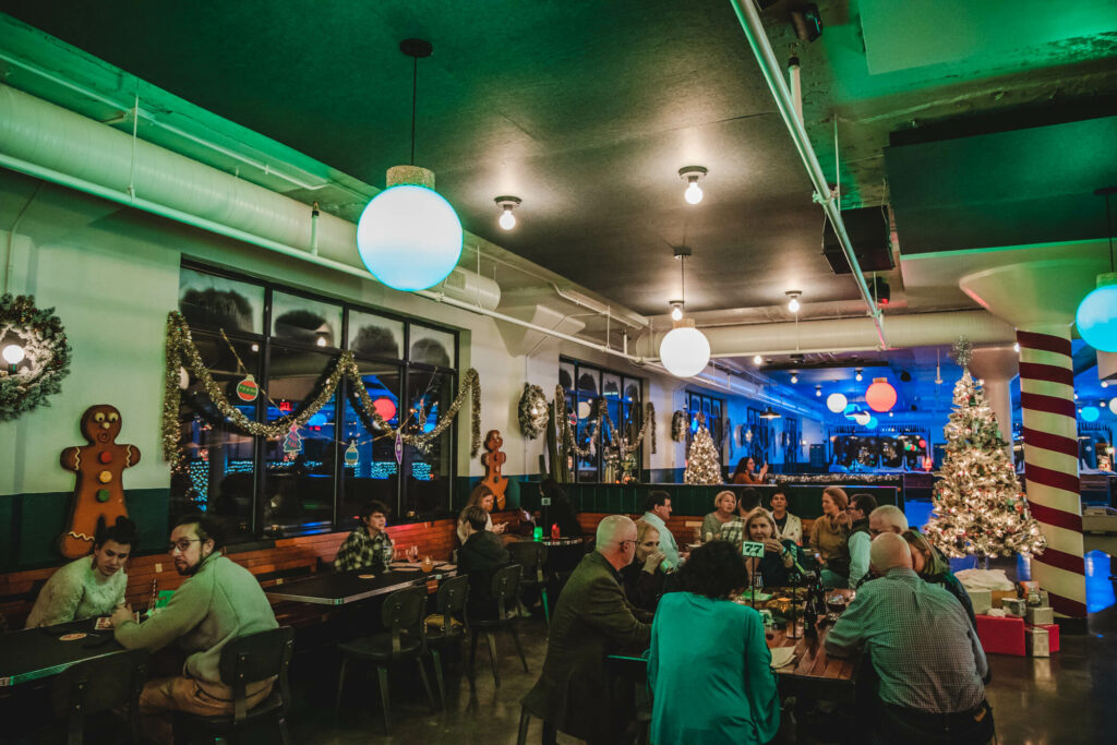 A festive restaurant setting with people sitting at tables, perfectly embodying "Deck the Halls." Holiday wreaths, garlands, gingerbread figures, and a lit Christmas tree adorn the room. Blue and green lighting cast a cozy glow, enhancing the inviting holiday ambiance.