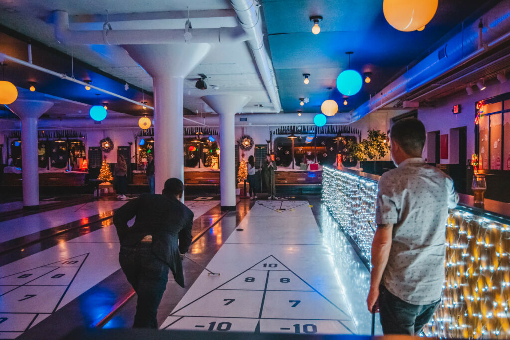 Two people playing shuffleboard indoors on the festive Rec Deck. One takes a shot while the other watches, surrounded by twinkling lights and colorful hanging lamps, creating a lively atmosphere.