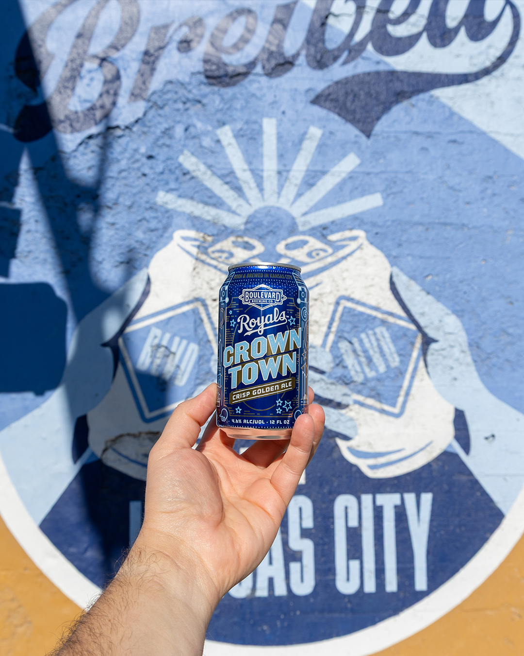 A hand holds a blue can of Boulevard Brewing Co.'s "Crown Town" Crisp Golden Ale. In the background, a blue mural with the words "Brewed" and "Kansas City" is partially visible.