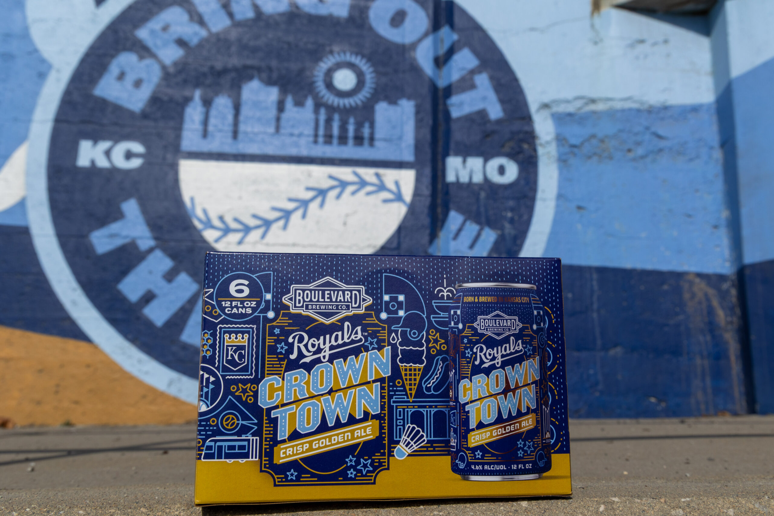 A blue and yellow six-pack of Boulevard Royals Crown Town Crisp Golden Ale rests in front of a sports-themed mural. The mural features a baseball and the words "Bring Out," with "KC MO" and skyline graphics.