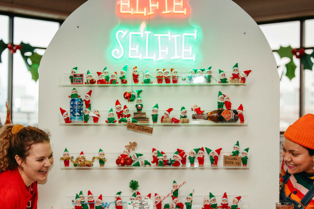 Two people smiling in front of a display filled with toy elves in playful poses. A neon sign above reads "Elfie Selfie." The Rec Deck backdrop includes holiday decorations like holly leaves and red bows, perfectly capturing the spirit of decking the halls.