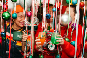 Three people are celebrating at a festive party on the rec deck, holding colorful drinks. They're surrounded by ornaments, ribbons, and holiday decorations. The scene is cheerful and vibrant as they truly deck the halls with joy.