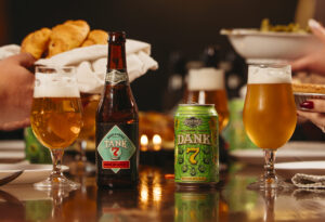 Two beers in glasses, one next to a bottle with a label reading "Tank 7", the other sitting next to a can with a label reading "Dank 7". In the background hands pass plates of food back and forth.