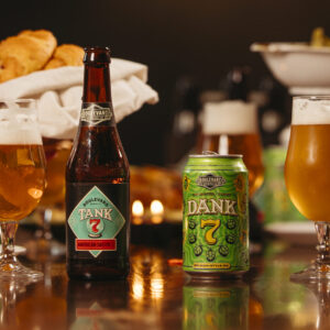 Two beers in glasses, one next to a bottle with a label reading "Tank 7", the other sitting next to a can with a label reading "Dank 7". In the background hands pass plates of food back and forth.