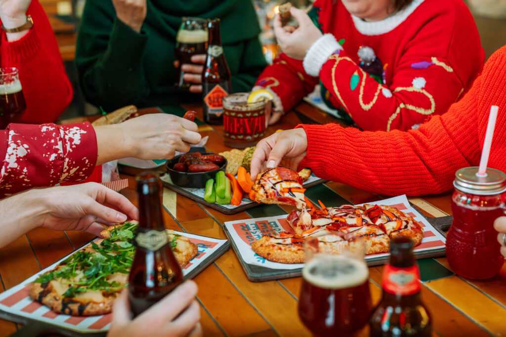 A group of people enjoying a meal at a wooden table, sharing pizza, flatbread, and drinks. Dressed in festive sweaters, they toast with glasses amidst the lively atmosphere. It's a Rec Deck the Halls gathering where laughter flows as freely as the beverages.