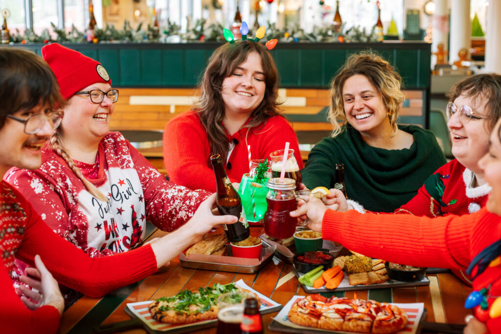 A joyful group in festive attire gathers around a table filled with holiday-themed delights, clinking bottles in celebration as "Deck the Halls" plays softly, enhancing the cheerful and decorated setting.