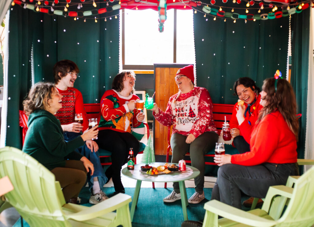 A group of people in festive sweaters are joyfully gathered around a table, sharing drinks and snacks. Seated on colorful chairs under twinkling string lights, with a curtain backdrop decorated with small lights, they rec Deck the Halls in a cozy holiday atmosphere.