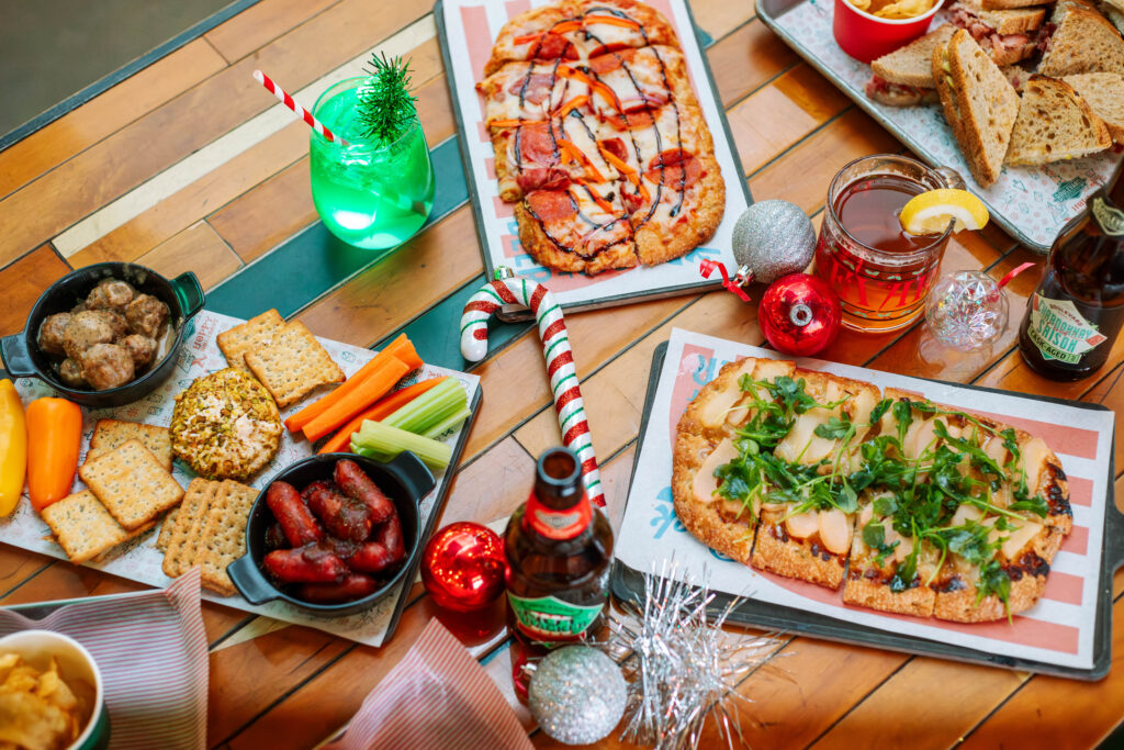 A festive table spread with various appetizers, like pizzas garnished with greens and meats, is reminiscent of a “Deck the Halls” celebration. Crackers, cheese, sausages, and meatballs sit alongside holiday-themed drinks and decorations, with colorful ornaments adding a cheerful touch.