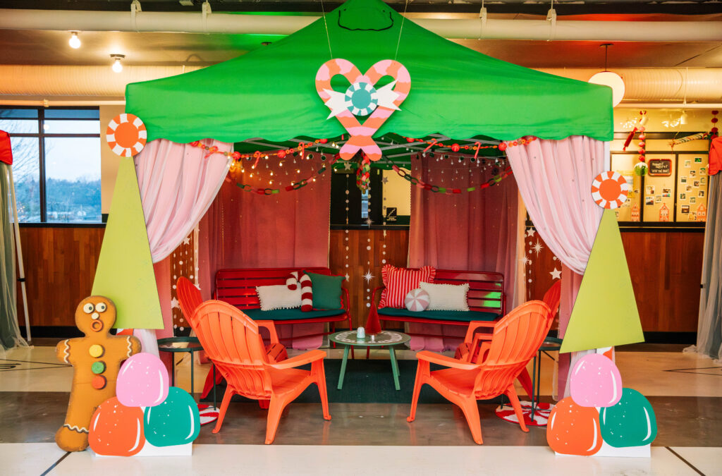 A festive tent decorated with a green roof and pink curtains, exuding a 'Rec Deck the Halls' charm with its candy-themed ornaments. Inside, red benches with pillows surround a glass table and orange chairs. A gingerbread man and oversized pastel candies welcome guests at the entrance.