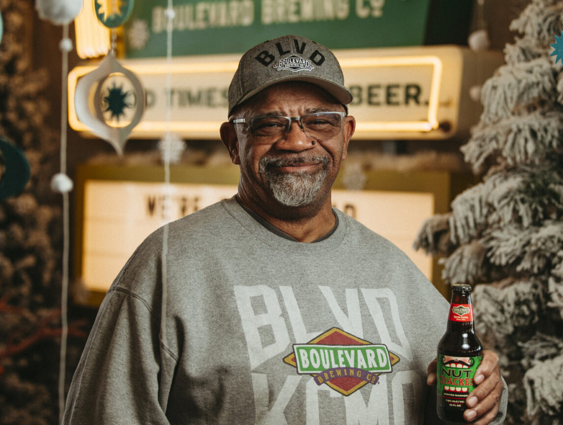 Byron Ewing in a "Boulevard Brewing" sweatshirt and cap holds a bottle of Nutcracker Ale, standing in front of a festive holiday display with a snowy tree and neon signs.