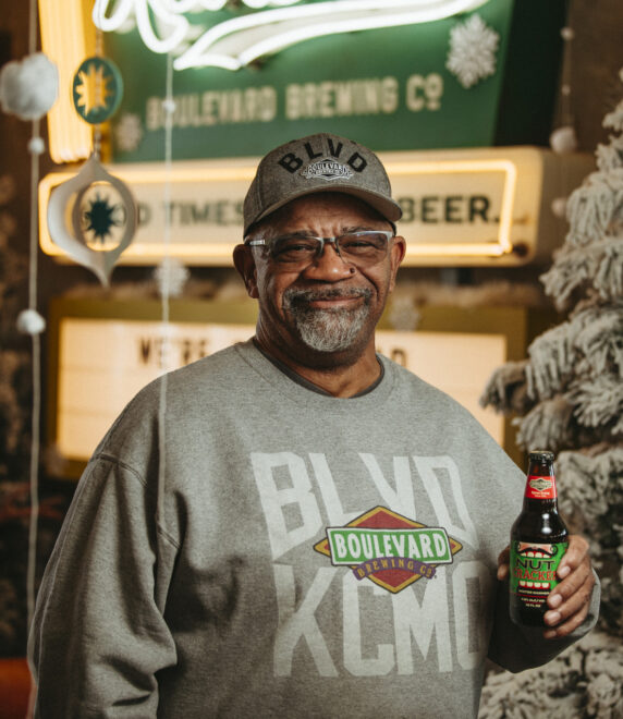 Byron Ewing in a "Boulevard Brewing" sweatshirt and cap holds a bottle of Nutcracker Ale, standing in front of a festive holiday display with a snowy tree and neon signs.