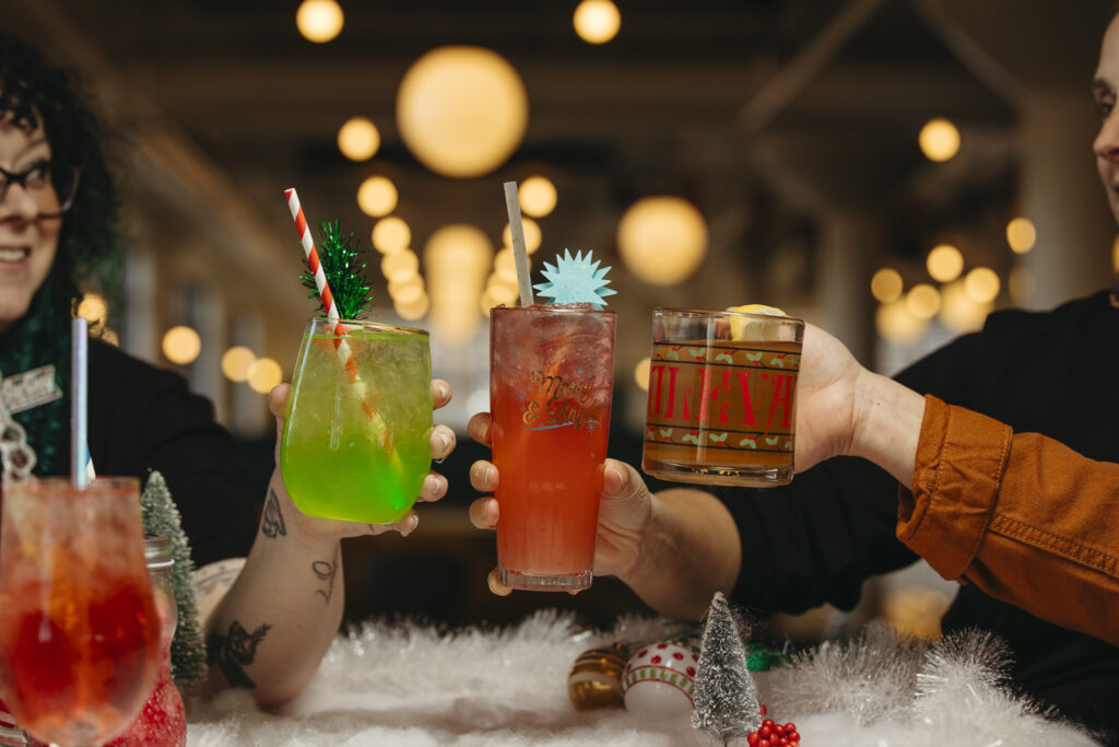 Three people holding festive cocktails on the Rec Deck in a holiday-themed setting. One drink is green with a red straw and decoration, another is red with a blue stirrer, and the last is amber with a lemon slice. Blurred lights glow in the background.