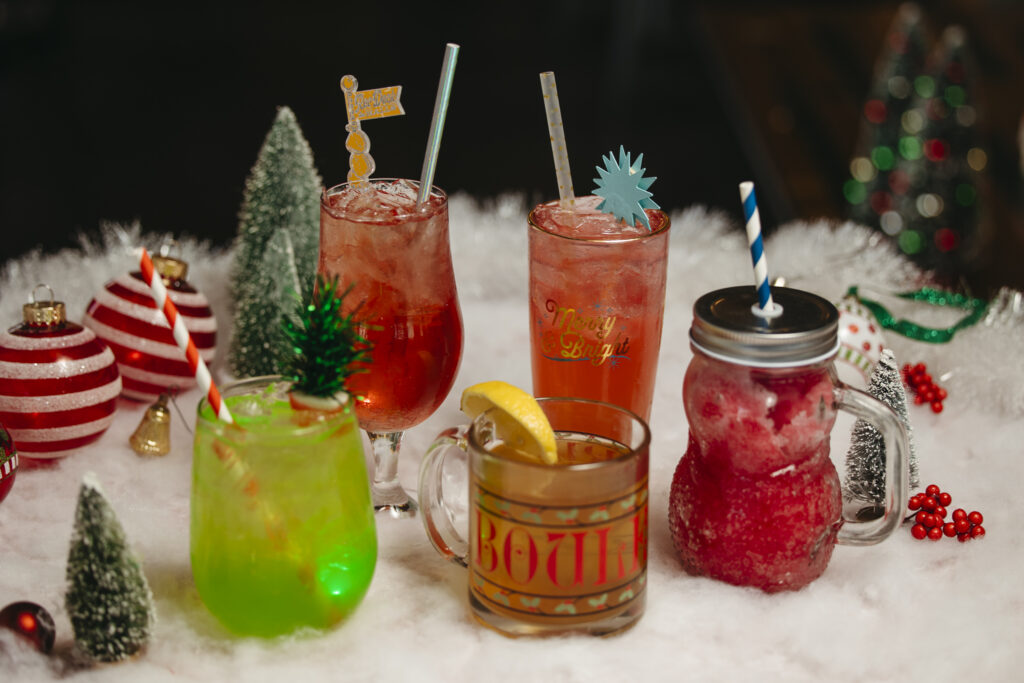 Festive holiday-themed drinks on a snowy table, featuring colorful cocktails with decorative stirrers and straws. Surrounding the drinks are Christmas ornaments, miniature trees, and berry accents, creating a cheerful "Deck the Halls" vibe in this vibrant atmosphere.