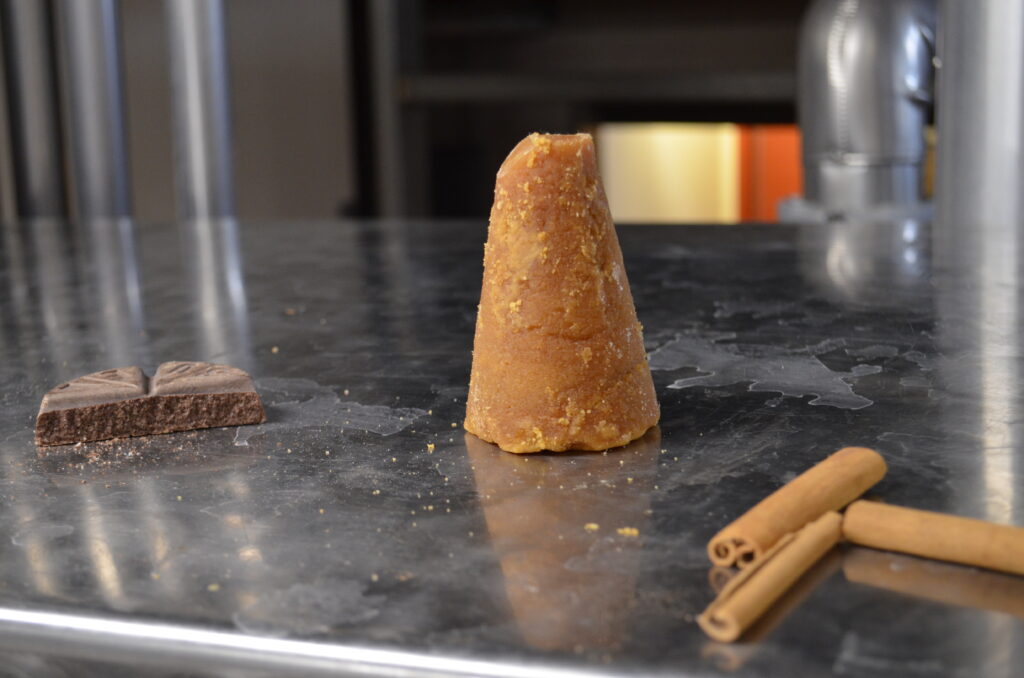 A cone-shaped piece of brown sugar (piloncillo) stands on a shiny metal surface, surrounded by a bar of chocolate and several cinnamon sticks. The background is slightly blurred, suggesting an indoor setting.