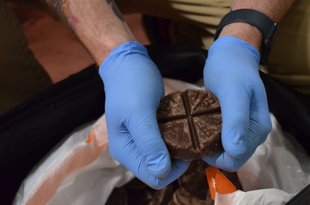 Person wearing blue gloves holding a round, segmented chocolate disc over a bag filled with similar discs.