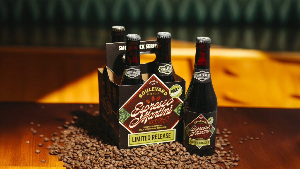 A four-pack of Boulevard Brewing Co.'s Espresso Martini Stout is displayed on a wooden table surrounded by coffee beans. The bottles are dark with labels matching the box, which reads "Limited Release" and features coffee-themed graphics.