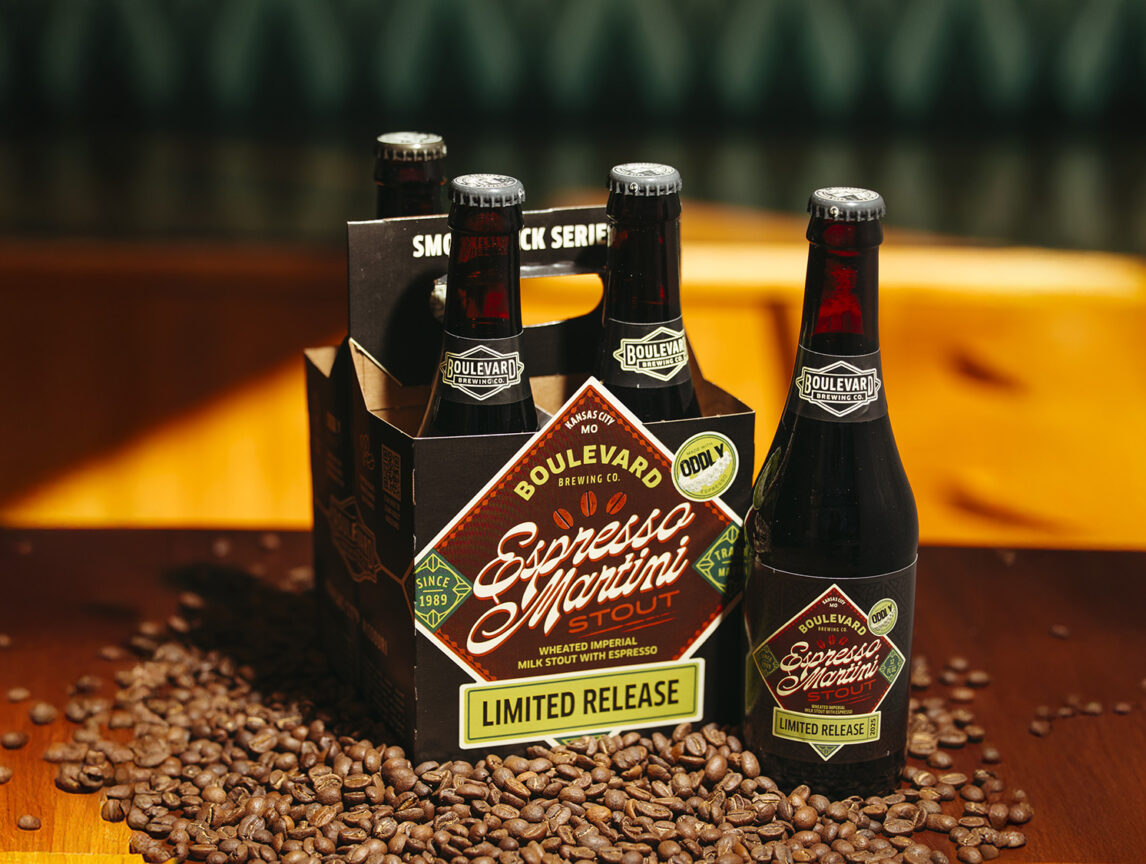 A four-pack of Boulevard Brewing Co.'s Espresso Martini Stout is displayed on a wooden table surrounded by coffee beans. The bottles are dark with labels matching the box, which reads "Limited Release" and features coffee-themed graphics.