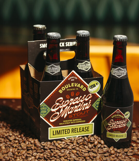 A four-pack of Boulevard Brewing Co.'s Espresso Martini Stout is displayed on a wooden table surrounded by coffee beans. The bottles are dark with labels matching the box, which reads "Limited Release" and features coffee-themed graphics.