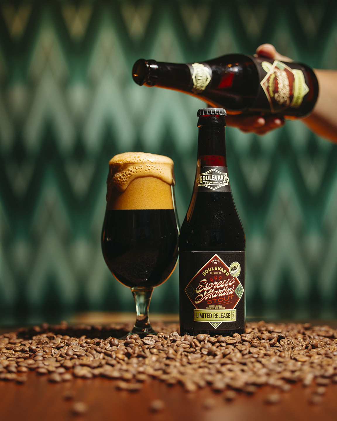 A bottle of Boulevard Brewing Co.'s limited release beer is being poured into a glass, with a frothy head overflowing. The glass stands on a table covered with coffee beans, against a blurred, patterned background.