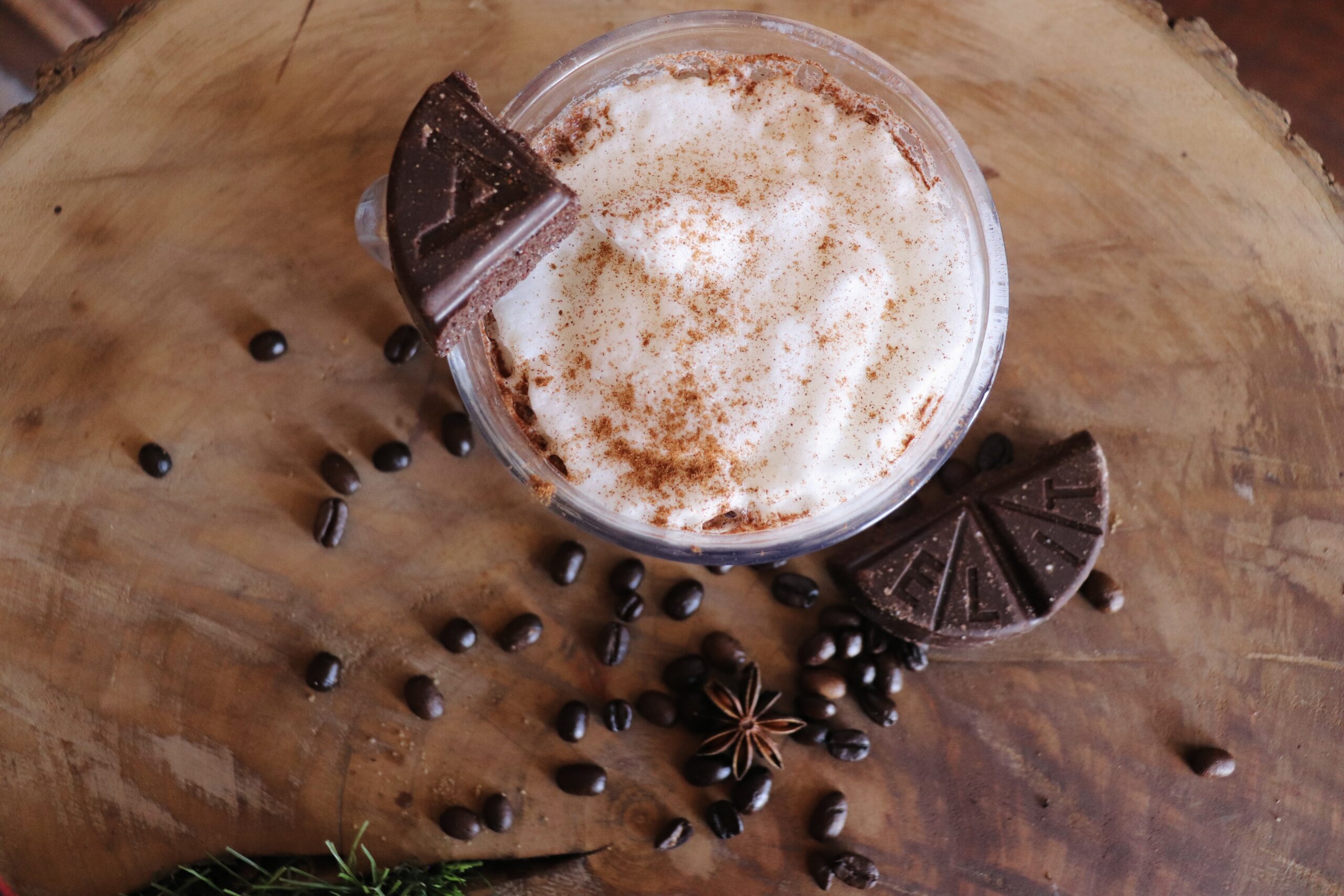 A creamy coffee drink topped with whipped cream and cinnamon, served in a glass mug. Garnished with triangular pieces of dark chocolate and surrounded by coffee beans, anise stars, and greenery on a wooden surface.