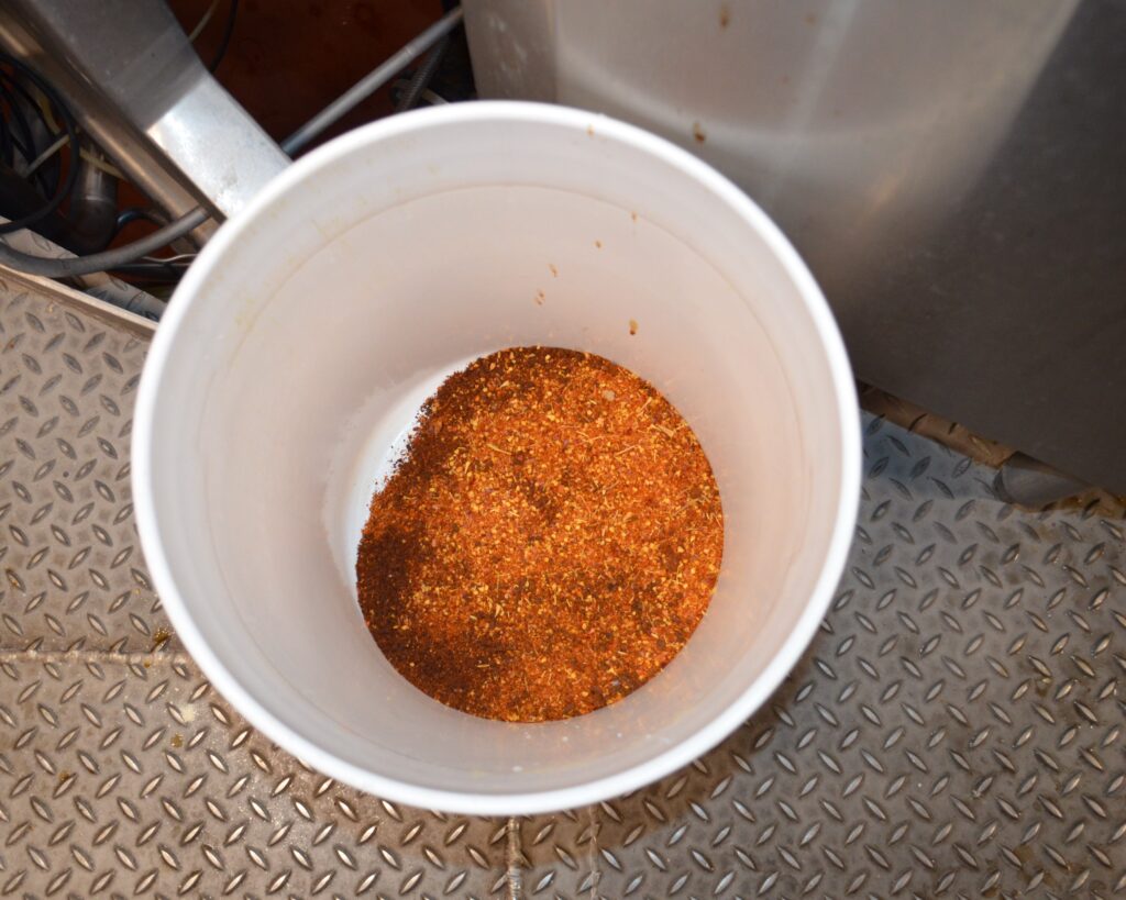 A white bucket filled with a reddish-brown spice mix sits on a textured metal floor.