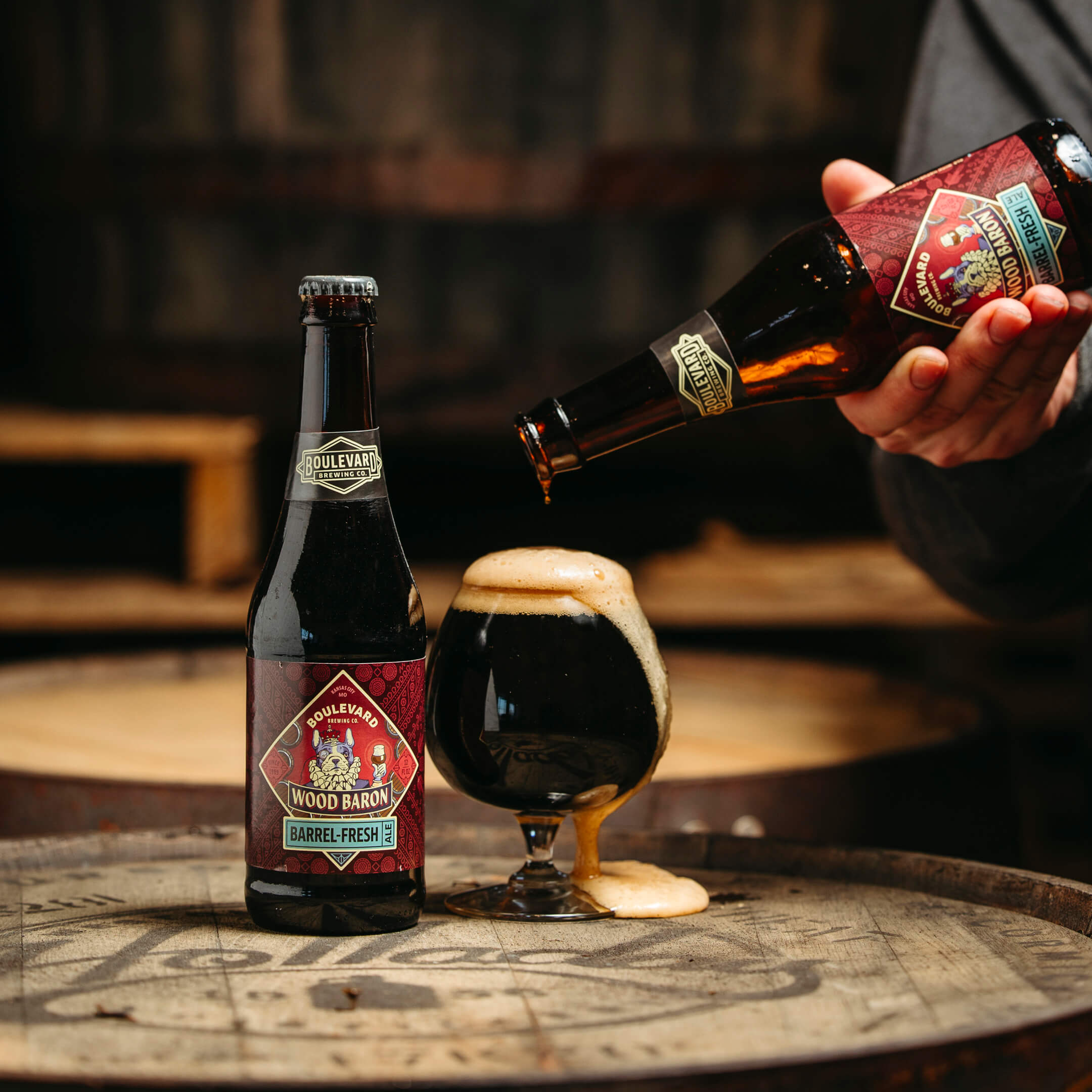 Two bottles of Boulevard Brewing Company's Wood Baron beer are shown. One bottle is being poured into a glass filled with dark beer on a wooden barrel surface. The label features a red design with text "Barrel Fresh.