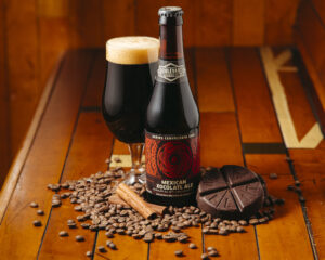 A bottle of Mexican Xocolatl Ale sits on a wooden table surrounded by coffee beans, chocolate, and a cinnamon stick. Next to the bottle is a glass filled with dark ale.