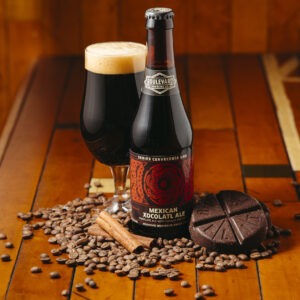 A bottle of Mexican Xocolatl Ale sits on a wooden table surrounded by coffee beans, chocolate, and a cinnamon stick. Next to the bottle is a glass filled with dark ale.