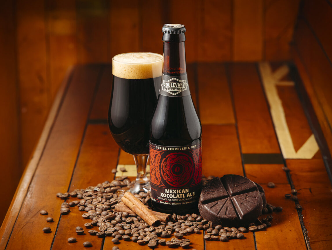 A bottle and glass of Mexican Xocolatl Ale sit on a wooden surface, surrounded by cocoa beans, chocolate pieces, and cinnamon sticks, creating a warm and inviting atmosphere.