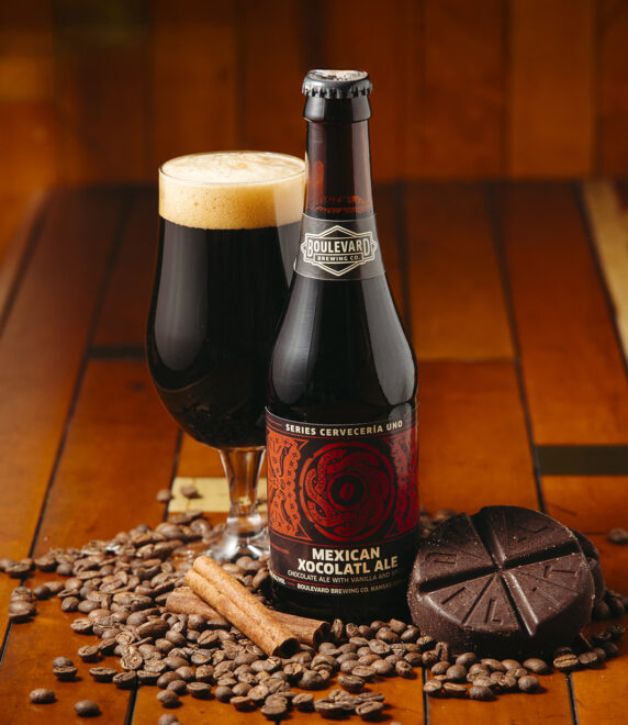 A bottle and glass of Mexican Xocolatl Ale sit on a wooden surface, surrounded by cocoa beans, chocolate pieces, and cinnamon sticks, creating a warm and inviting atmosphere.