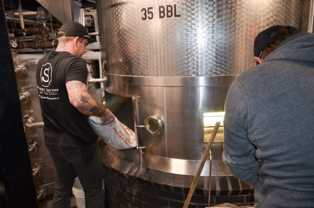 Two people work near a large stainless steel brewing tank labeled "35 BBL." One person is pouring ingredients, and the other is tending to the tank. Pipes and equipment surround them in the brewery.