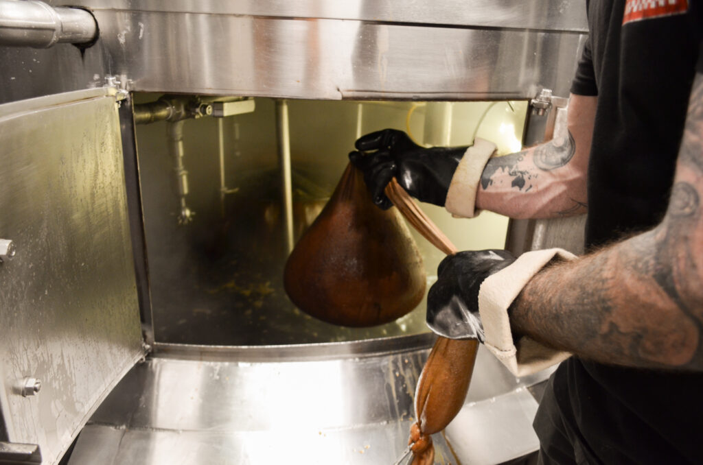 In the pristine, metallic brewhouse, a tattooed worker with gloves holds a dark-colored sack connected to machinery, hinting at the intricate process of crafting drinks. Pipes and equipment gleam in this industrial setting designed for precision and quality.