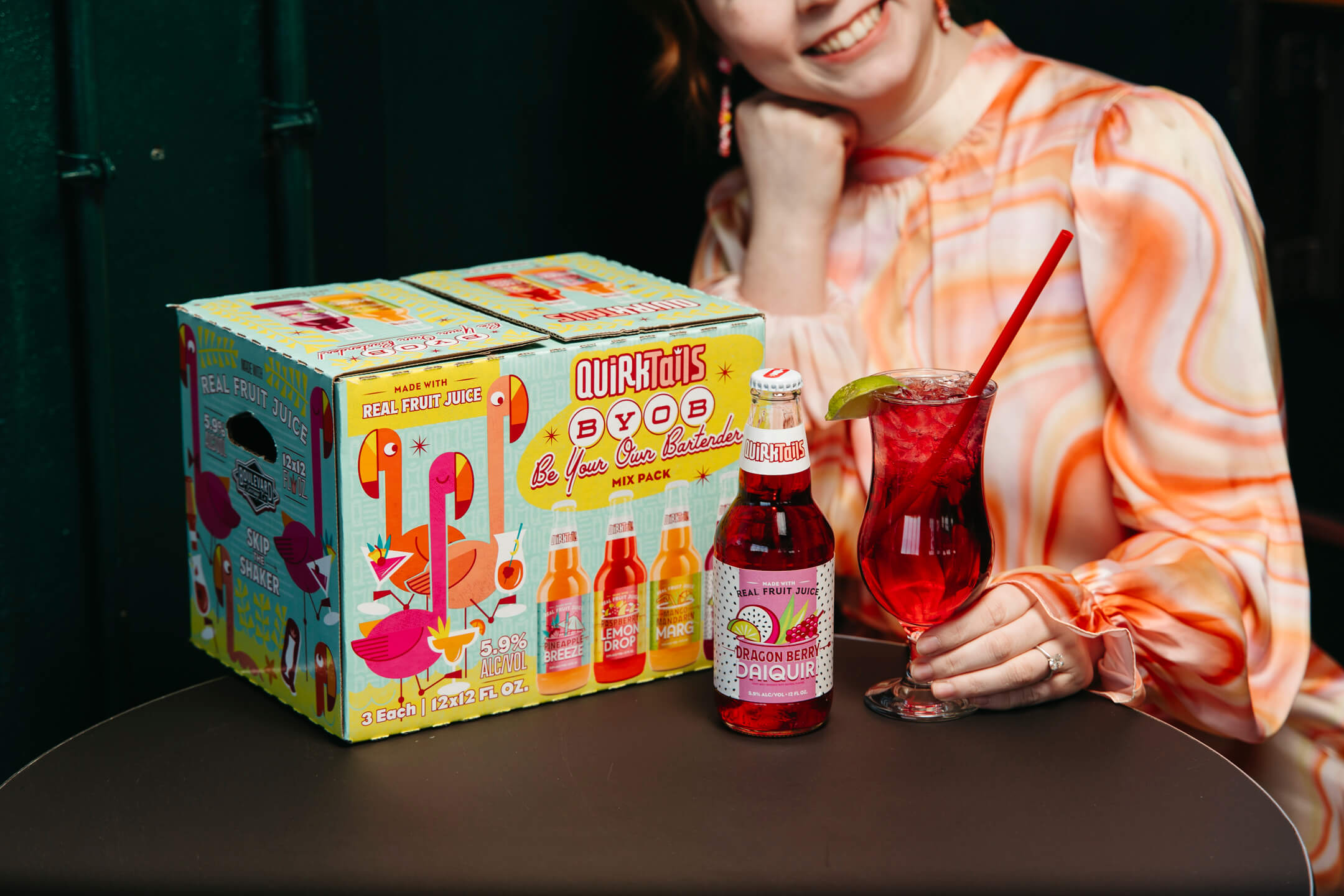 A person in a patterned blouse sits at a table with a boxed pack of Quirktails Horizon Spice Dragon Berry drinks. They hold a bottle next to a glass of red drink garnished with a lime and a red straw.